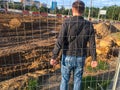 A young strong guy stands behind a metal fence and holds on to it. the guy climbed over the bars to get to the construction site,