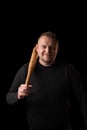 A young strong man holds a bat in his hand and smiles. Dark background. Man in black clothes. Bouncer, security guard, bodyguard