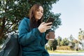 Young student freelancer woman in park sitting on bench, drinking coffee outdoor and using mobile cell phone smiling Royalty Free Stock Photo