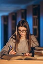 Young student in glasses preparing for the exam. Girl in the evening sits at a table in the library with a pile of books Royalty Free Stock Photo