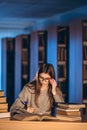 Young student in glasses preparing for the exam. Girl in the evening sits at a table in the library with a pile of books