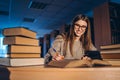 Young student in glasses preparing for the exam. Girl in the evening sits at a table in the library with a pile of books, smiling