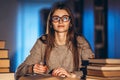 Young student in glasses preparing for the exam. Girl in the evening sits at a table in the library with a pile of books Royalty Free Stock Photo