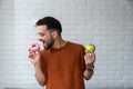 Young student man doubts what to choose healthy food or sweets junk unhealthy food holding green apple and donuts in hands