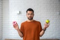 Young student man doubts what to choose healthy food or sweets junk unhealthy food holding green apple and donuts in hands