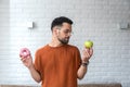 Young student man doubts what to choose healthy food or sweets junk unhealthy food holding green apple and donuts in hands