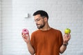 Young student man doubts what to choose healthy food or sweets junk unhealthy food holding green apple and donuts in hands