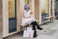 Young stylish Arabic woman in hijab drinking coffee on bench on city after shopping