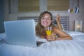 Beautiful Caucasian woman 30s lying on bed at night in home bedroom using internet at laptop computer drinking healthy orange juic Royalty Free Stock Photo