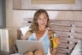 Beautiful Caucasian woman 30s sitting on bed at night in home bedroom using internet at laptop computer drinking healthy orange ju Royalty Free Stock Photo