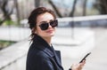 Young stylish beautiful brunette girl in sunglasses looking at camera holding her smartphone in street in spring