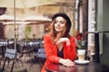 Young stylish beautiful woman sitting in city cafe in red jacket, street style, drinking aromatic coffee. Elegant girl in hat Royalty Free Stock Photo