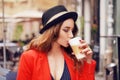 Young stylish beautiful woman sitting in city cafe in red jacket, street style, drinking aromatic coffee. Elegant girl in hat Royalty Free Stock Photo