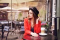 Young stylish beautiful woman sitting in city cafe in red jacket, street style, drinking aromatic coffee. Elegant girl in hat Royalty Free Stock Photo