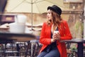 Young stylish beautiful woman sitting in city cafe in red jacket, street style, drinking aromatic coffee. Elegant girl in hat Royalty Free Stock Photo