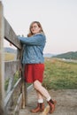 Young blonde woman in jeans jacket and red dress near old wooden fence on the mountains background Royalty Free Stock Photo