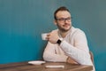 A young stylish brooding fellow businessman from Europe with glasses, with a beard and in a white shirt drinking coffee sitting at