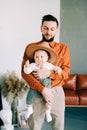 A young stylish father in trend orange shirt plays with his young son in a brown hat in his arms in a spacious stylish Royalty Free Stock Photo