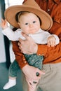 A young stylish father in trend orange shirt plays with his young son in a brown hat in his arms in a spacious stylish Royalty Free Stock Photo