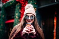 Young stylish girl in a burgundy coat and a knitted hat holds coffee and smiles. dressed in sunglasses smiling at the camera Royalty Free Stock Photo