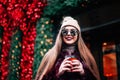 Young stylish girl in burgundy coat and knitted hat holds in hands coffee dressed in sunglasses smiling at camera Royalty Free Stock Photo