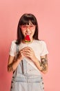 Young stylish girl in trendy sunglasses, with tattoos, posing in denim overalls, eating lollipop on pink