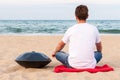 Young stylish guy sitting on the sand beach near handpan or hang with sea On Background. The Hang is traditional ethnic