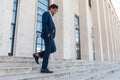 Young stylish man with hands in pockets walking down the stairs outside a white building. Wearing white shirt and dark blue suit Royalty Free Stock Photo