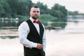 Young stylish man in a waistcoat, horizontal portrait of the groom, portrait on a background of nature, the river and the pier