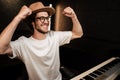 Young stylish musician happily raising hands up and showing power gesture during work in sound recording studio