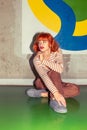 Young stylish redhead Caucasian woman in retro style of 70s sitting at wall vertical looking away