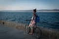 Young stylish woman in dress and with backpack is riding bicycle on promenade Royalty Free Stock Photo