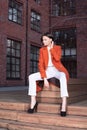 Young stylish woman in a red coat and white pants sitting on the wooden stairs