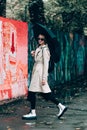 Young stylish woman wearing long beige coat, white boots, black hat, umbrella and backpack posing through the city streets.