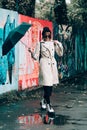 Young stylish woman wearing long beige coat, white boots, black hat, umbrella and backpack posing through the city streets.