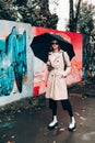 Young stylish woman wearing long beige coat, white boots, black hat, umbrella and backpack posing through the city streets.