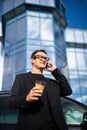 Young successful businessman wearing black suit and talking on his smartphone while standing near modern office or skyscrapers Royalty Free Stock Photo