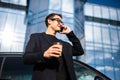 Young successful businessman wearing black suit and talking on his smartphone while standing near modern office or skyscrapers Royalty Free Stock Photo