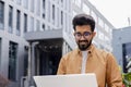 Young successful businessman working with laptop remotely sitting on bench outside office building, Hindu programmer in Royalty Free Stock Photo