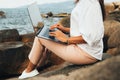 Young successful, smiling girl working using a laptop, sitting on a rocky seashore. traveling, blogger, freelancer, content plan,