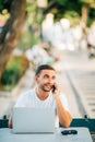 Young successful smiling smart man or student in casual shirt, glasses sitting at table, talking on mobile phone in city park usin Royalty Free Stock Photo