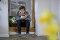 Young suffering guy with scarf having man flu, sitting in a door frame while blowing in his tea, holding a thermometer in his hand Royalty Free Stock Photo