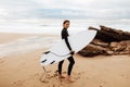 Young surfer lady walking with board on the sandy beach, looking and smiling at camera, full length shot Royalty Free Stock Photo