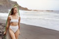 Young surfer woman in bikini going surfing stands with surfboard on the black sandy beach. Girl walks with longboard Royalty Free Stock Photo