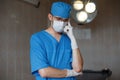 Young surgeon in a blue medical uniform in a protective white mask wearing rubber gloves is standing in the hospital and thinking