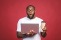 Young surprised african man standing and using laptop computer isolated over red background. Drinking coffee or tea Royalty Free Stock Photo
