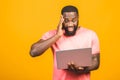 Young surprised african man standing and using laptop computer isolated over yellow background
