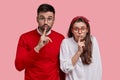 Young surprised couple make silence sign, stand closely to each other, keep index fingers on lips, demonstrate shush