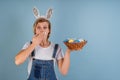 Young surprised woman with bunny ears putting her hand to the mouth holding a basket with Easter colored eggs  on blue Royalty Free Stock Photo