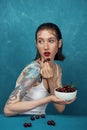 Young surprised woman sits at the table with cherry bowl in front of blue background